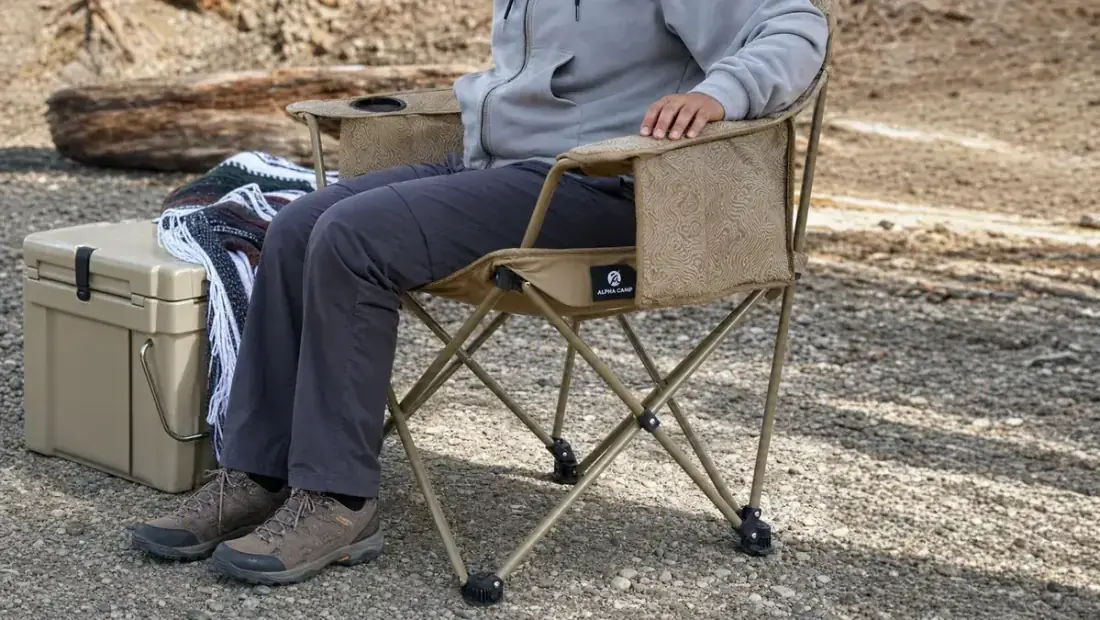 A man's hand gripping the padded armrest of the ALPHA CAMP oversized camping chair while lowering himself into it beside a cooler