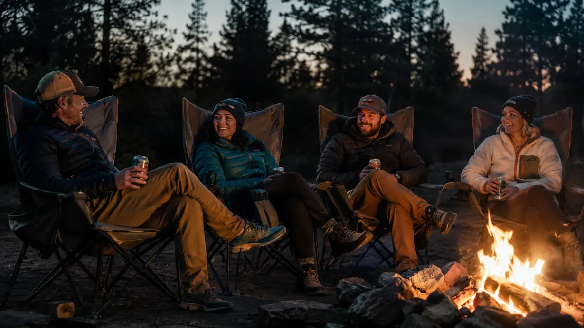 A small group of adults relaxing in oversized camping chairs around a campfire at dusk