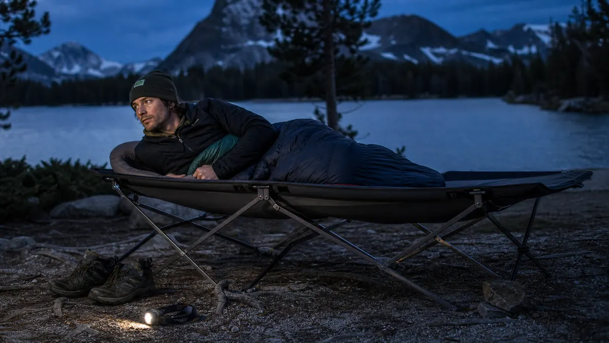 Folding camping cot sitting slightly uneven on a rocky, root-lined campsite pad at dusk