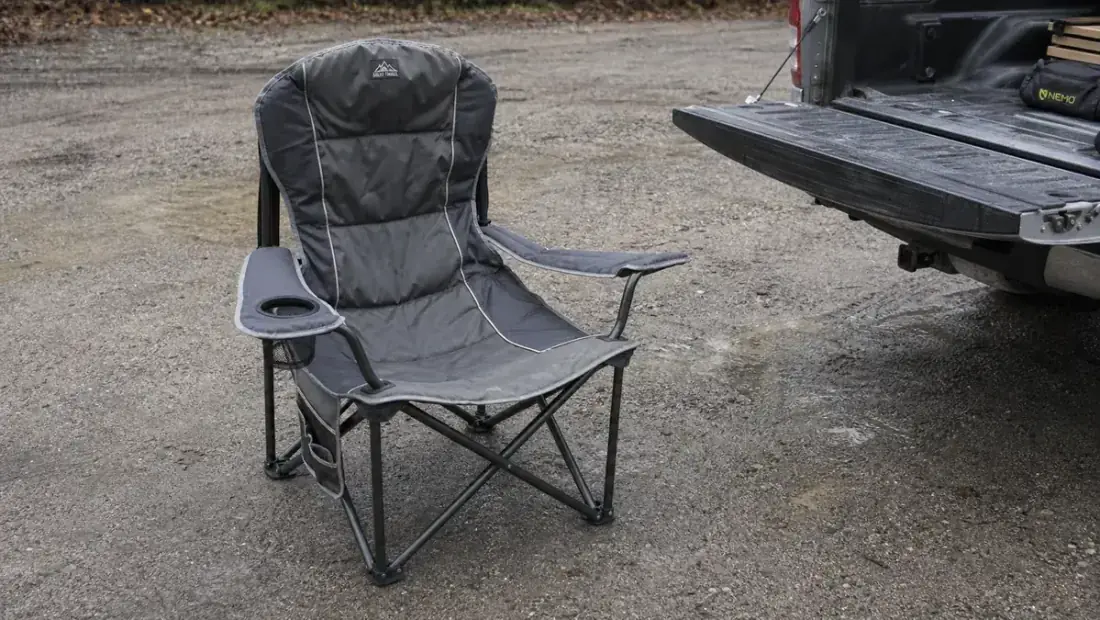 ALPHA CAMP oversized camping chair sitting empty in a gravel parking lot next to a truck tailgate, overcast light