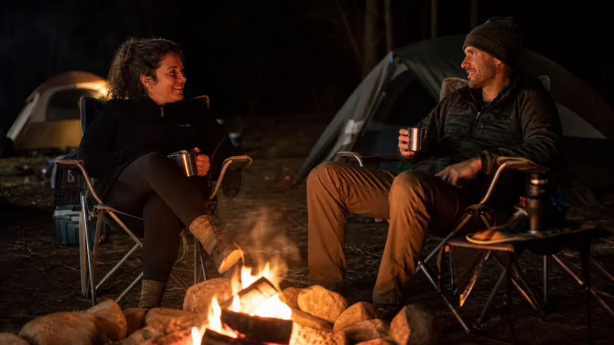 Two adults sitting in camping chairs around a fire at night with a family tent visible behind them