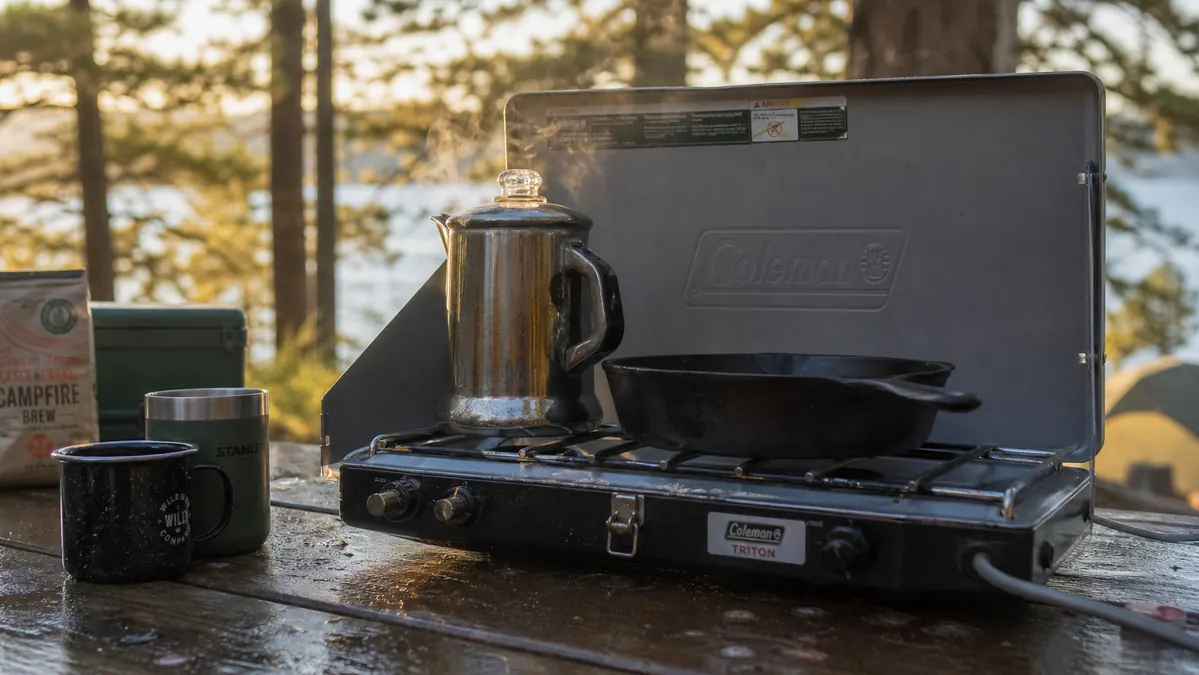 Two-burner propane camp stove sitting open on a picnic table at a campsite in early morning light, coffee pot on one burner