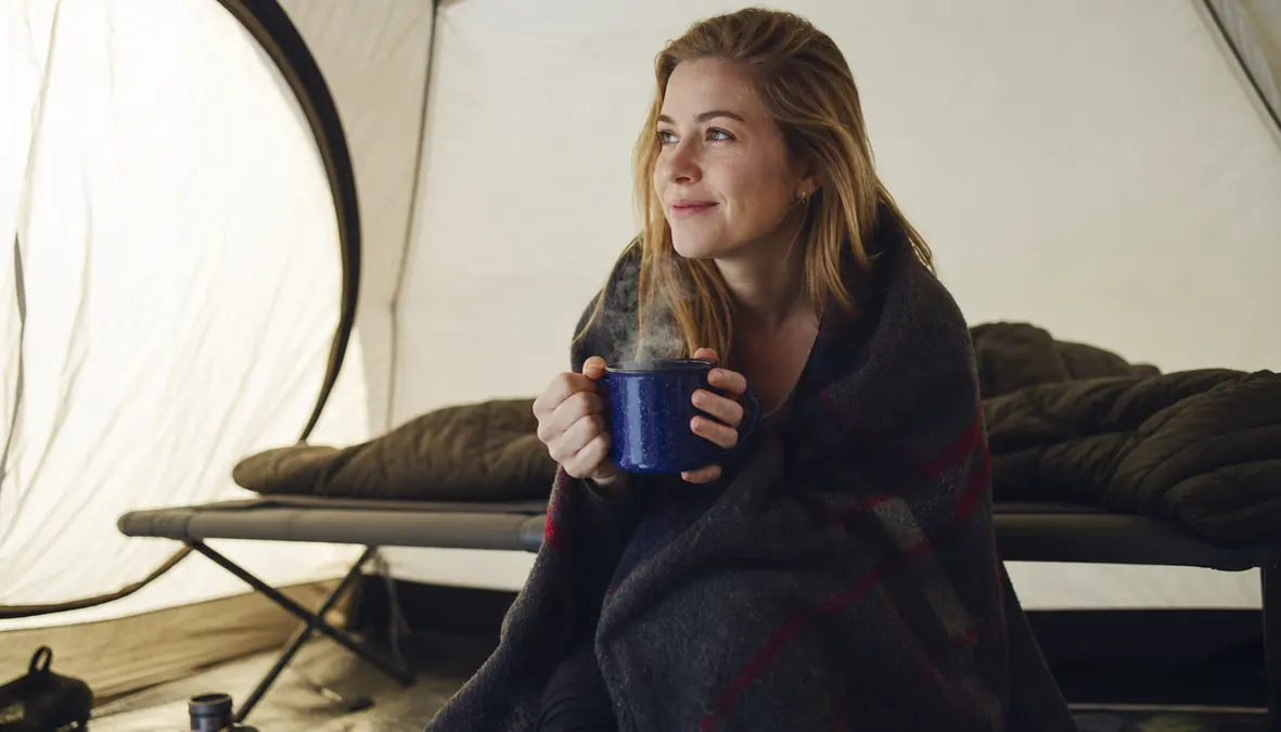 A camper sitting up on a camping cot inside a tent in the morning, wrapped in a wool blanket with a warm drink