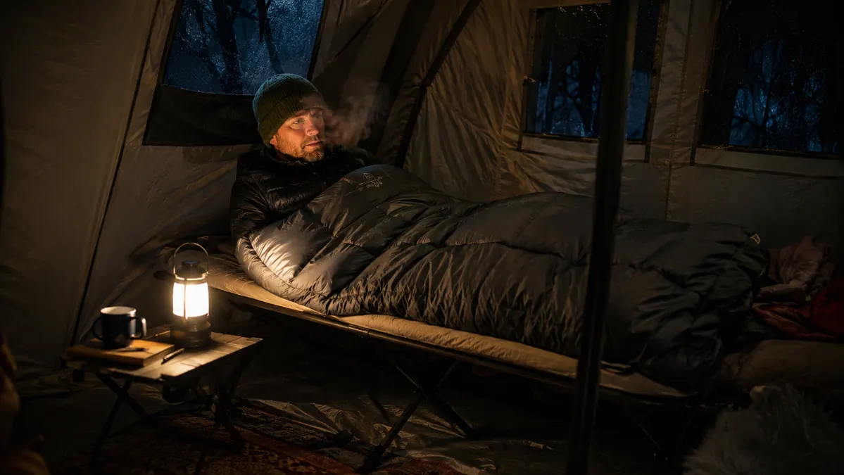 A camper bundled in a sleeping bag on a folding camp cot inside a tent at night, breath visible in the cold air